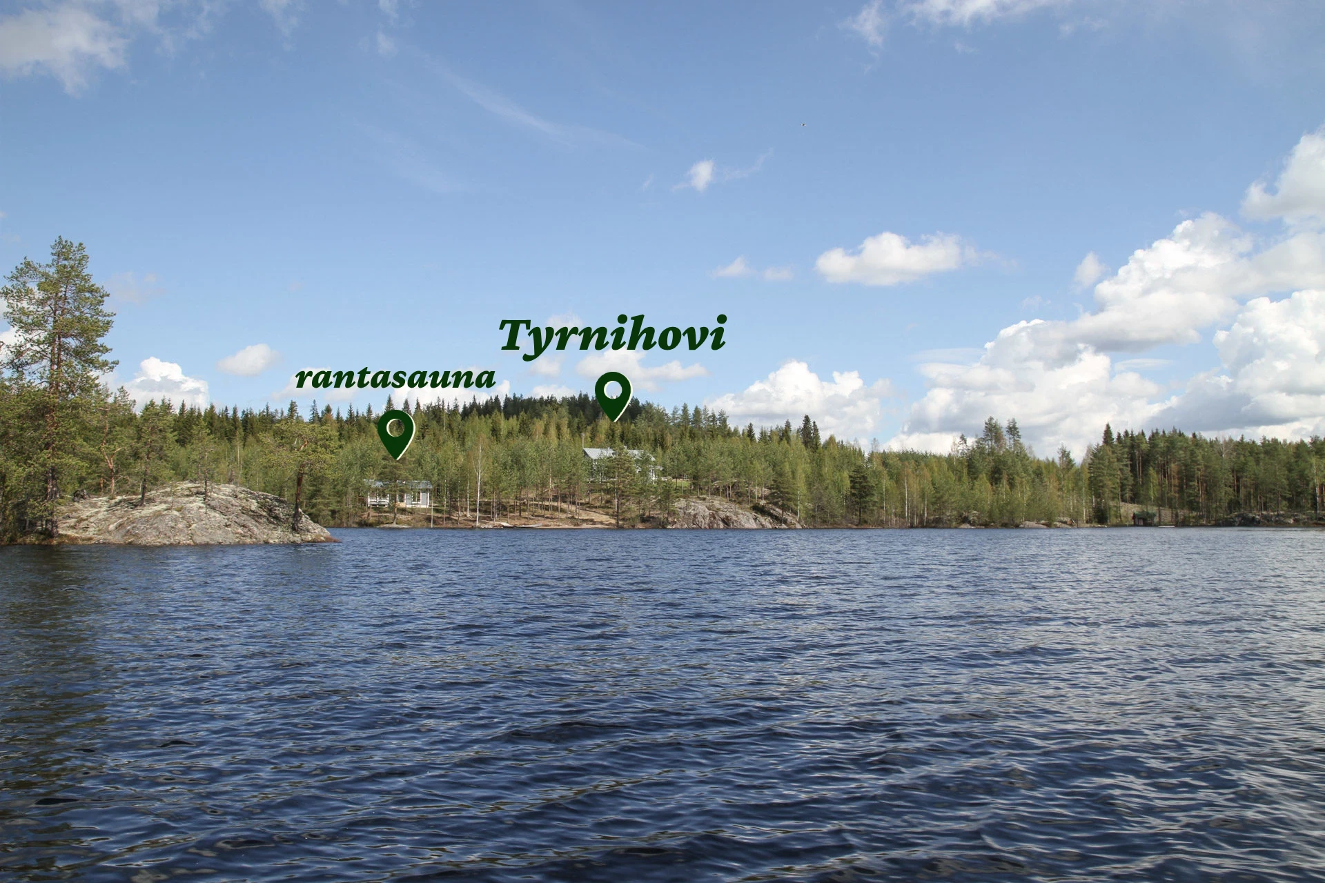 Näkymä Kallavedeltä kohti rantasaunaa ja Tyrnihovia, jotka sijaitsevat kauniisti kalliorinteellä metsän ympäröimänä. Sininen järvenselkä ja pilvinen taivas täydentävät luonnonrauhan tunnelman.